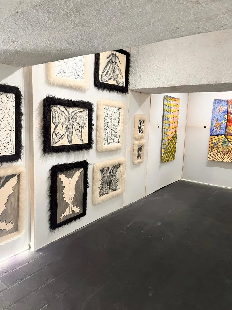 Desenhos emoldurados em pelúcia preta e creme, instalados em painéis de um núcleo expositivo, com peças em tecido pintado ao fundo